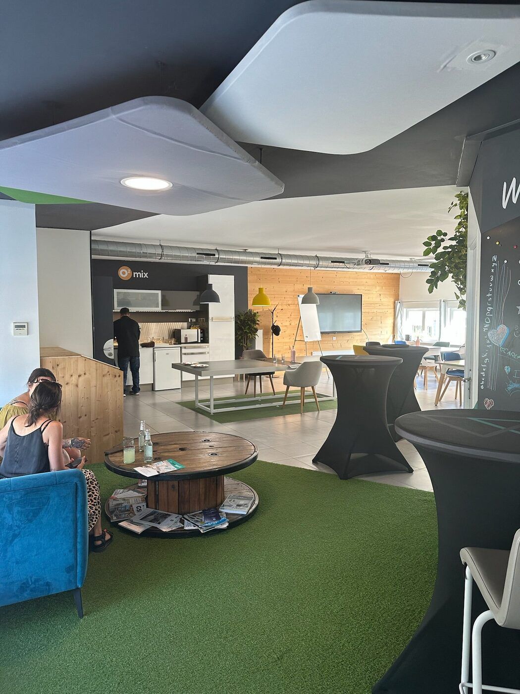 L'espace de vie d'Oxywork : pelouse synthétique, fauteuil bleu, table basse en touret, mange-debout et cuisine ouverte au fond