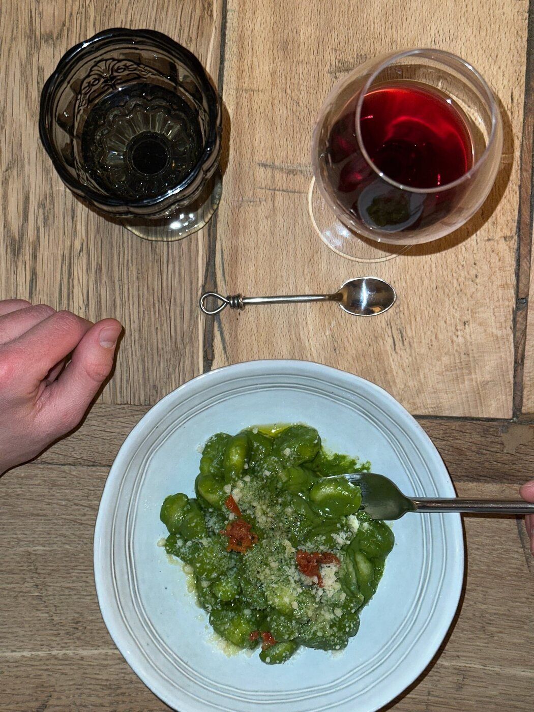 Vue de dessus d'une table en bois : une assiette de gnocchi au pesto et parmesan, un verre de vin rouge et les mains de deux convives